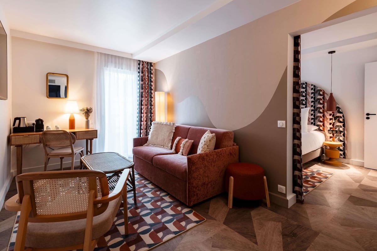 A hotel room with a sofa, a desk with a lamp, a chair, and a patterned rug.