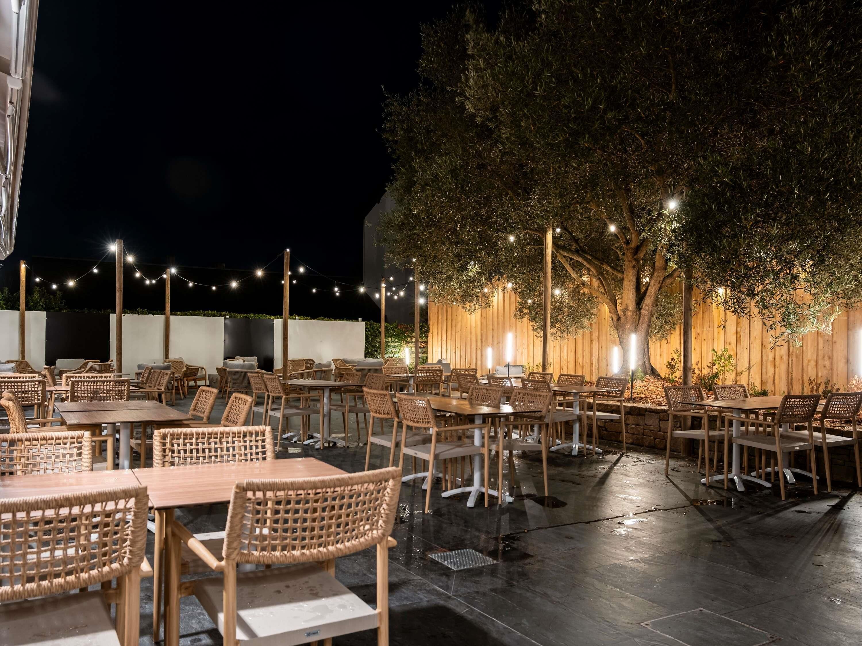 Un área de comedor al aire libre con mesas y sillas de mimbre, rodeada por una cerca de madera y luces colgantes.