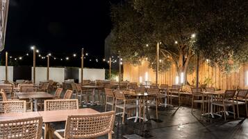 Un área de comedor al aire libre con mesas y sillas de mimbre, rodeada por una cerca de madera y luces colgantes.