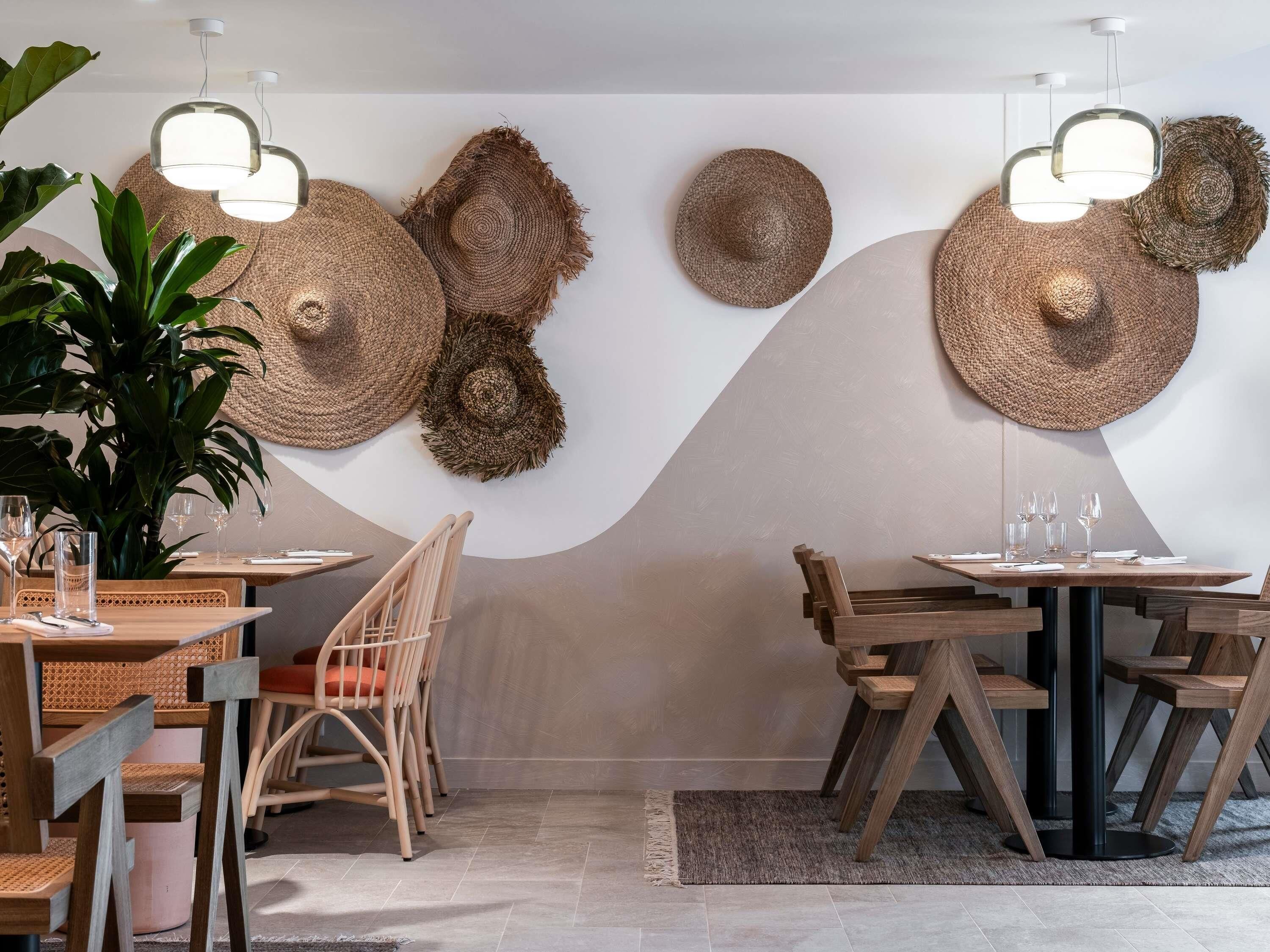 Un comedor con dos mesas de madera, sillas de mimbre y sombreros tejidos en la pared.