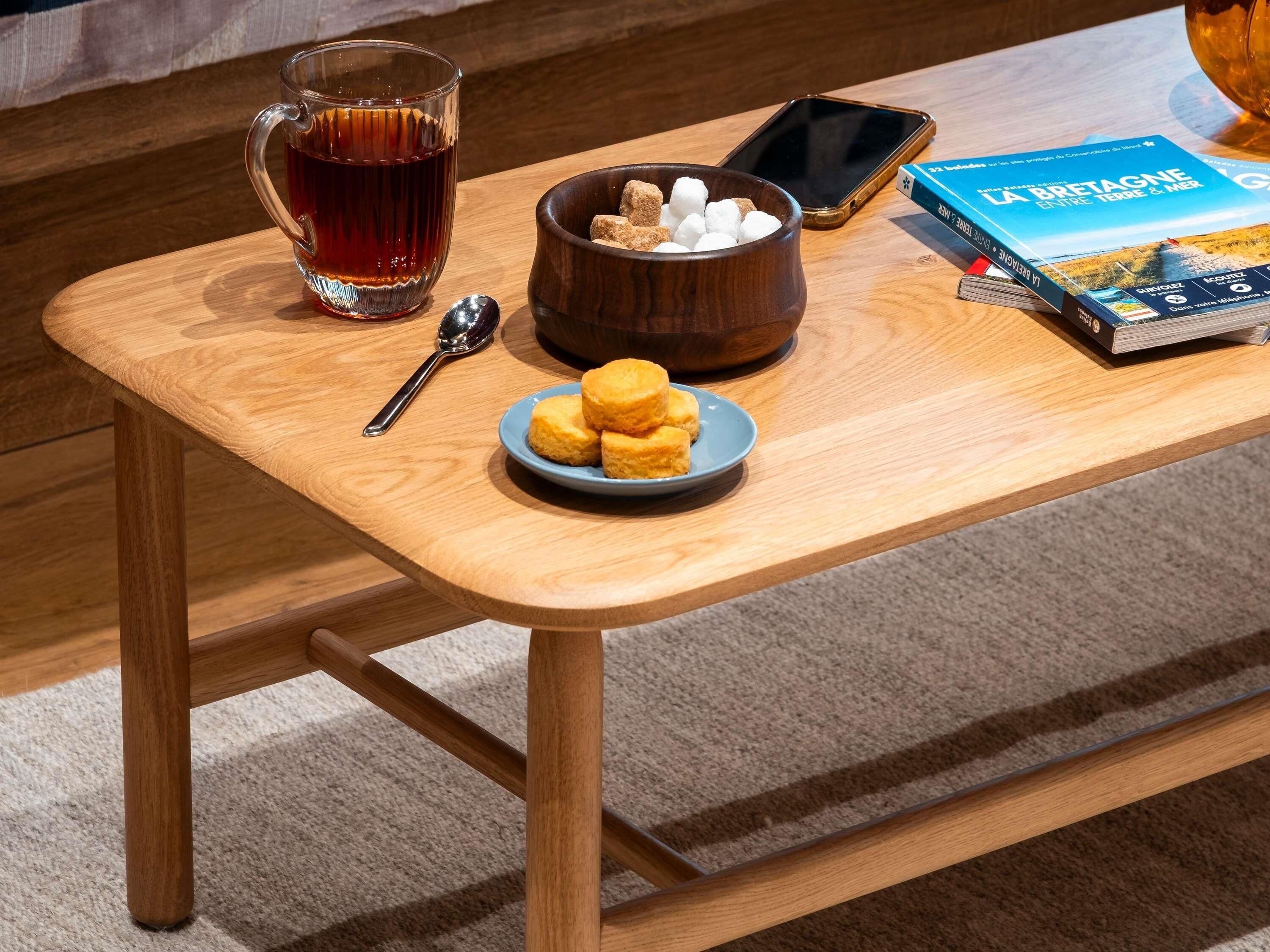 A wooden table with a glass of tea, a bowl of marshmallows, a spoon, a smartphone, and a book.