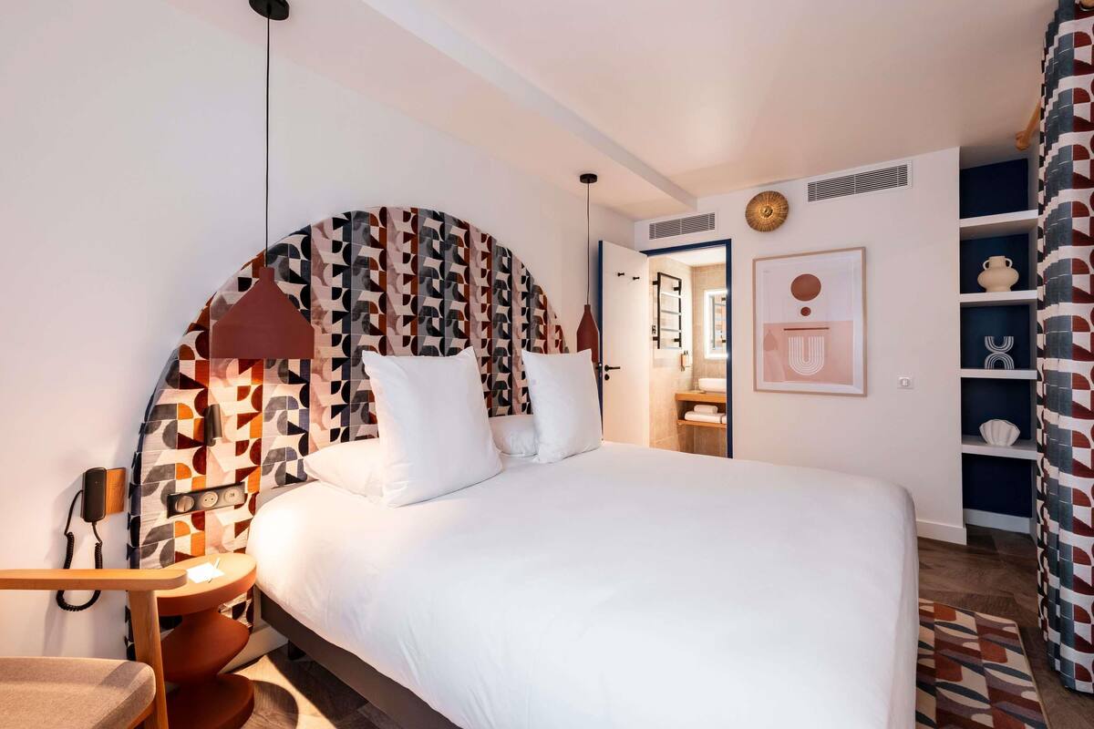 A modern bedroom with a large bed, a patterned headboard, a bedside table with a lamp, and a view into the bathroom.