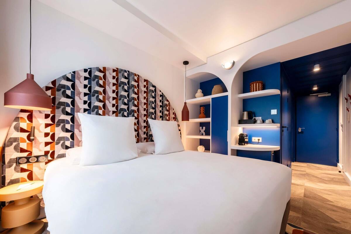 A modern hotel room with a large bed, a patterned headboard, built-in shelving, and a blue accent wall.