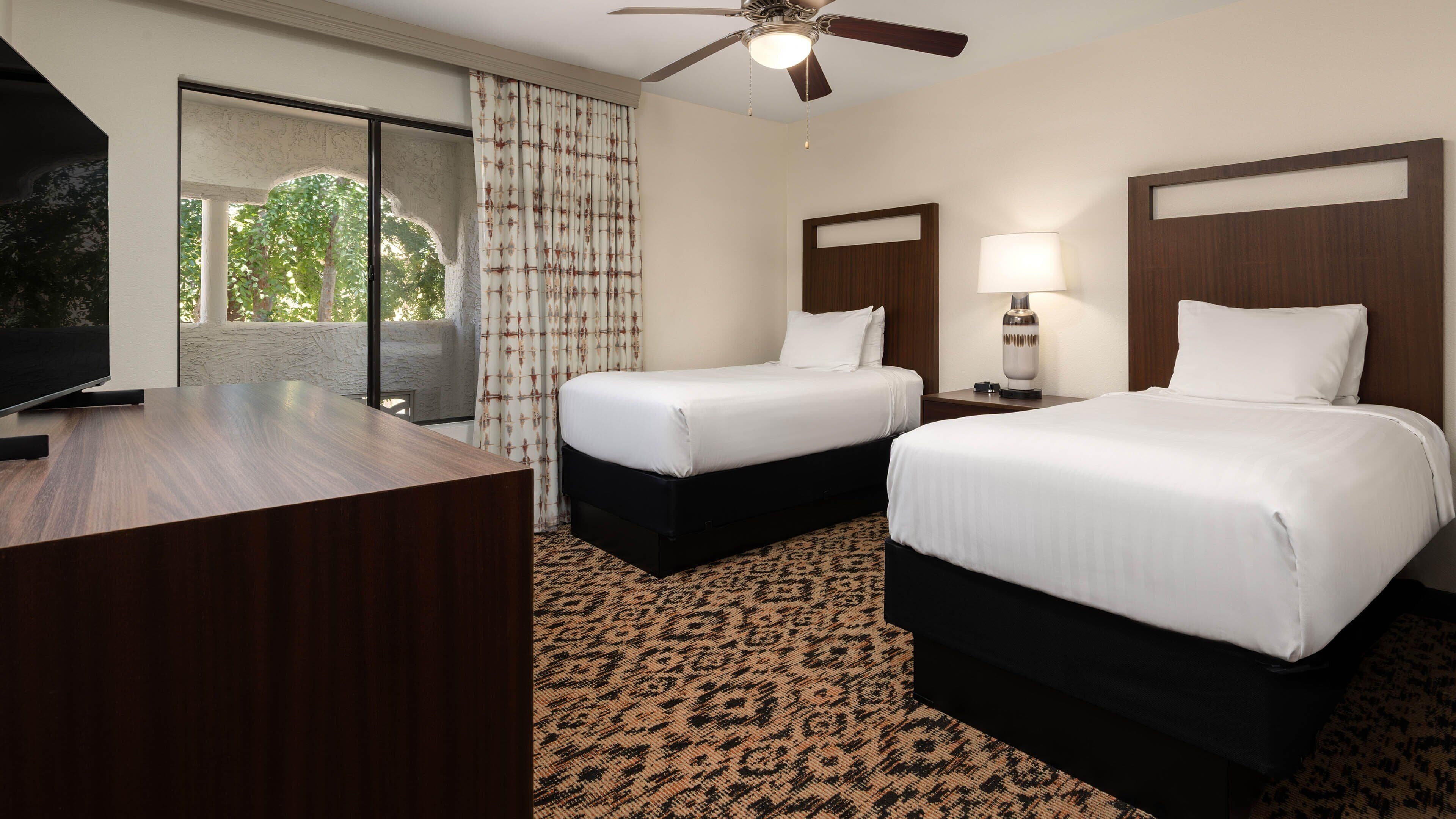 A hotel room with two beds, a desk, a ceiling fan, and a view of greenery outside.