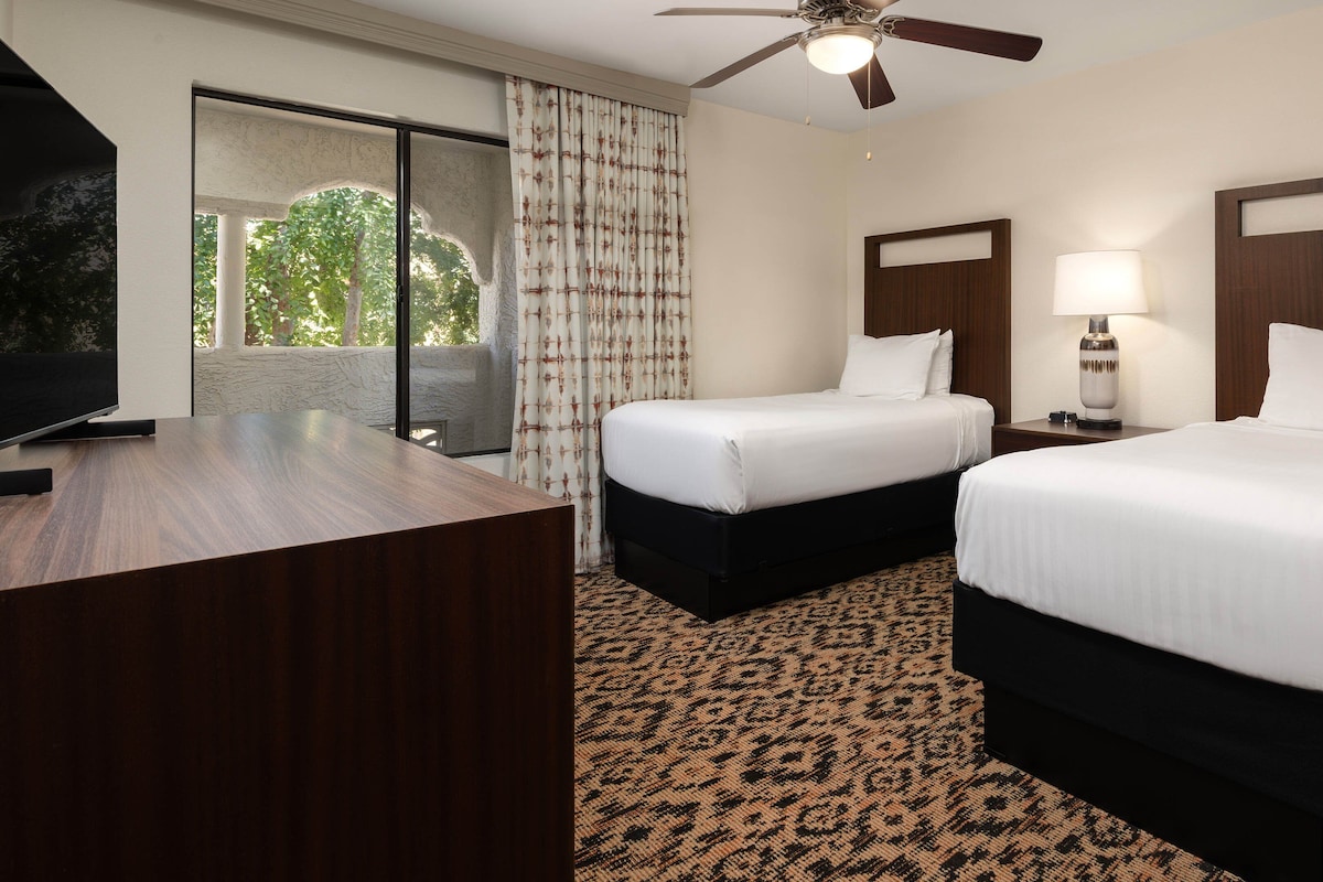 Une chambre d’hôtel avec deux lits, un bureau, un ventilateur de plafond et une vue sur la verdure environnante.