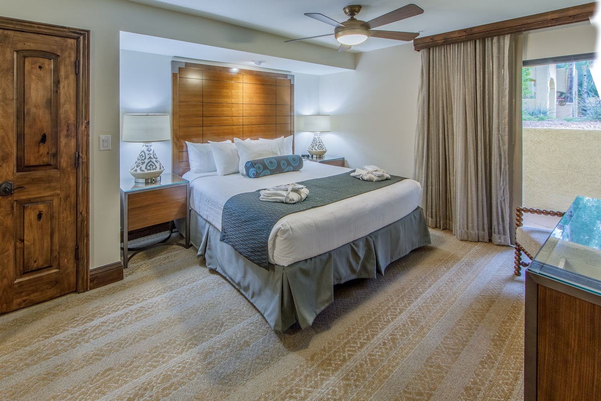 A hotel room with a large bed, wooden headboard, two bedside tables with lamps, a ceiling fan, and a view of the outdoors through a window.