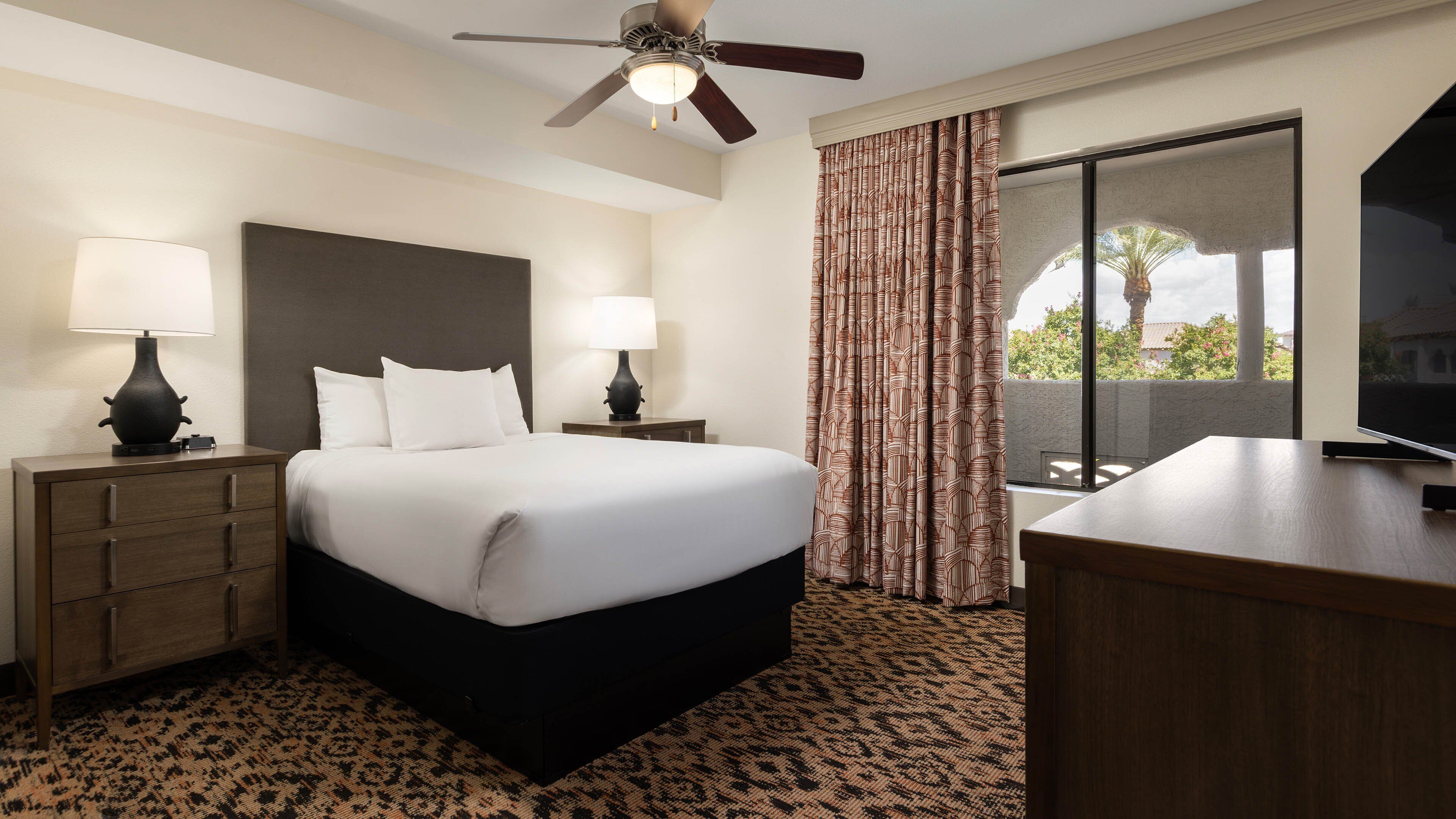 A hotel room with a bed, two bedside tables with lamps, a ceiling fan, a large window with curtains, and a TV on a wooden stand.