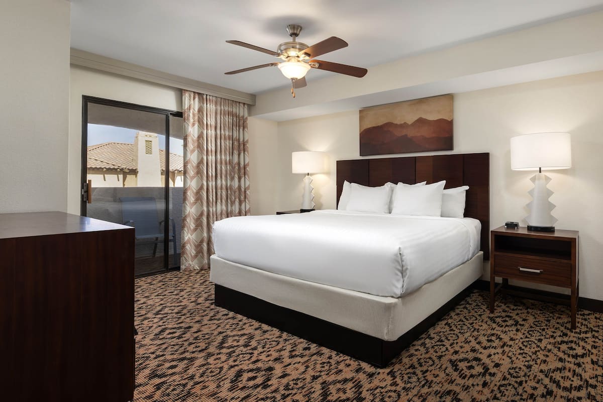 A hotel room with a large bed, a desk, two bedside lamps, a ceiling fan, and a window with curtains.