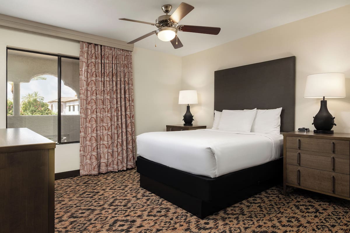 A hotel room with a bed, a desk, a television, a ceiling fan, and a view of the outside.