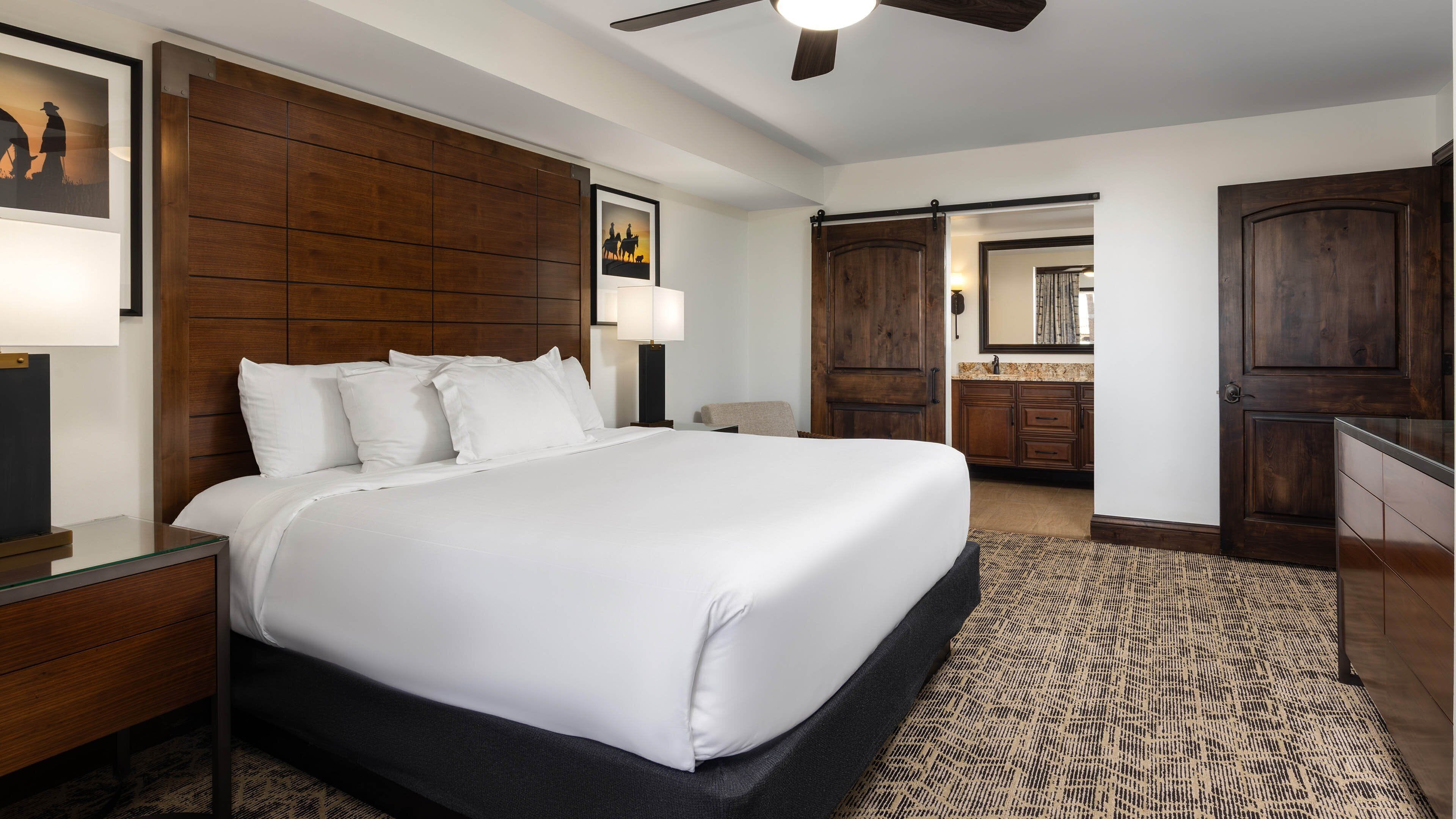 Ein Hotelzimmer mit einem großen Bett, einem Holzkopfteil, einem Deckenventilator, einem Badezimmer mit Spiegel und einer Tür zu einem weiteren Raum.