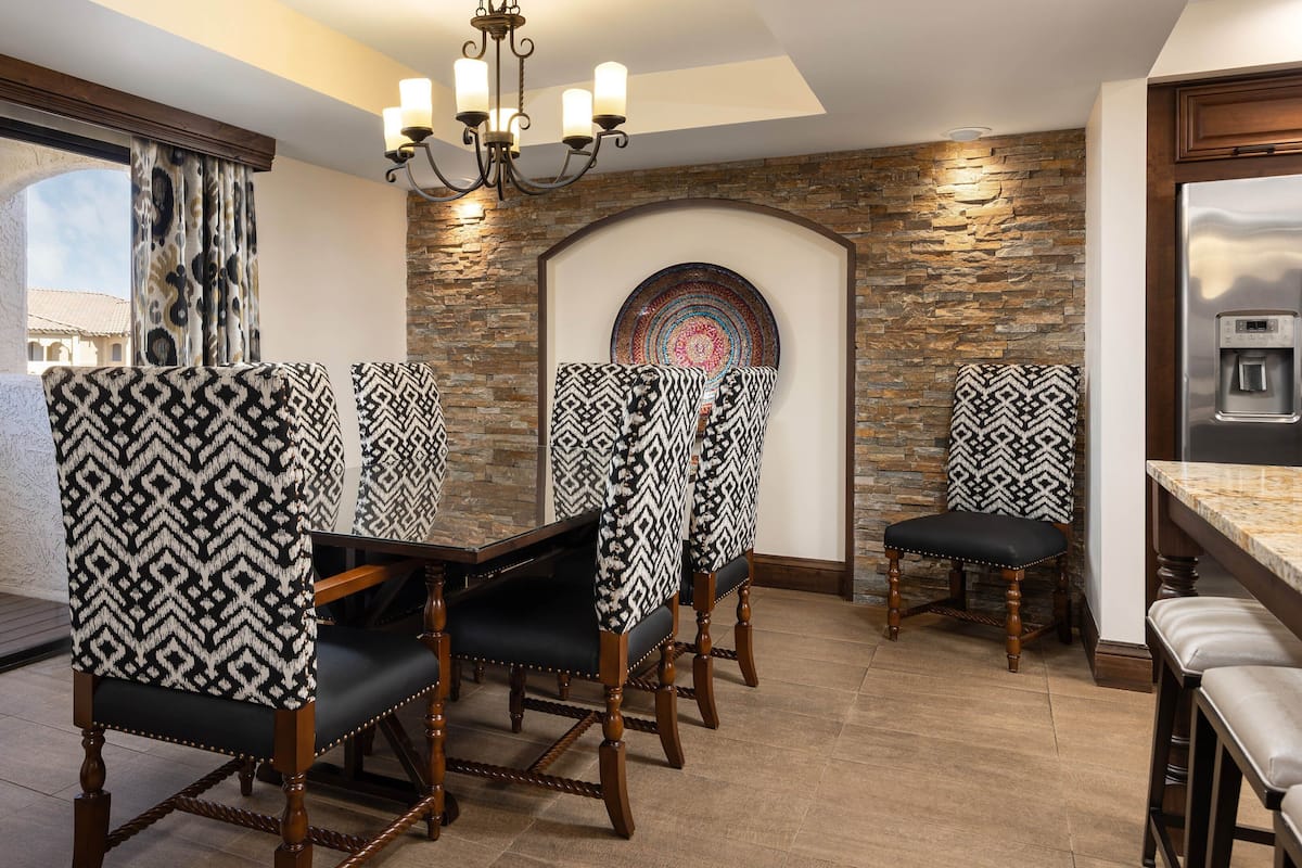 A dining area with patterned chairs, a chandelier, a stone wall, and a view of the outside.