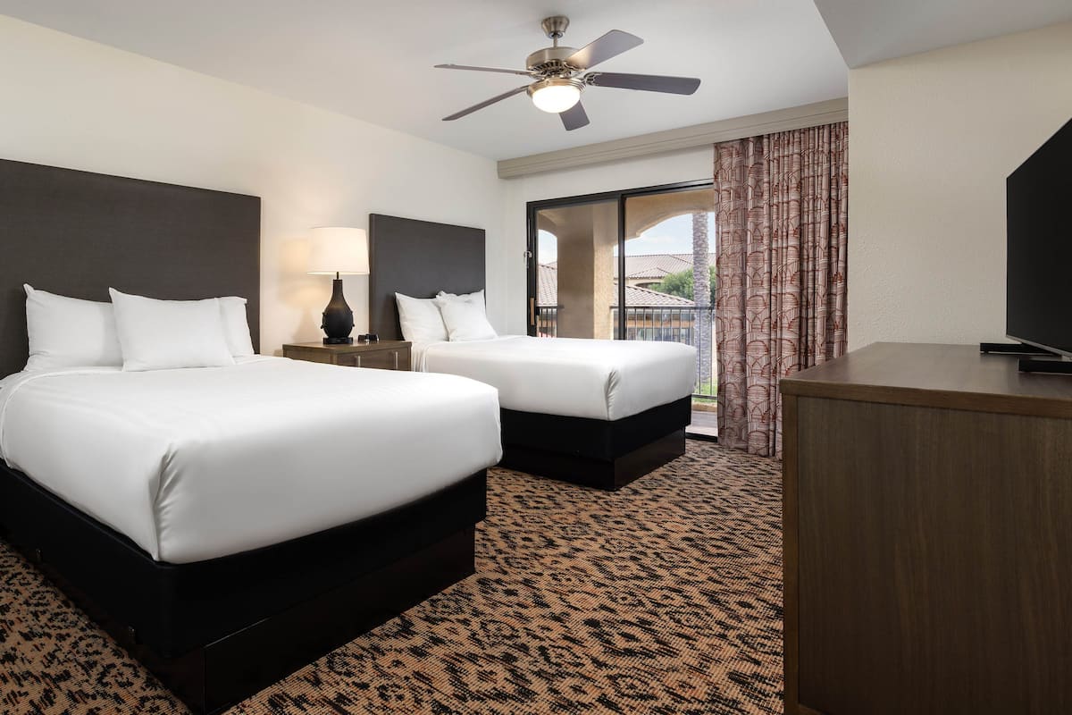 A hotel room with two beds, a ceiling fan, a TV, and a view of the outside.