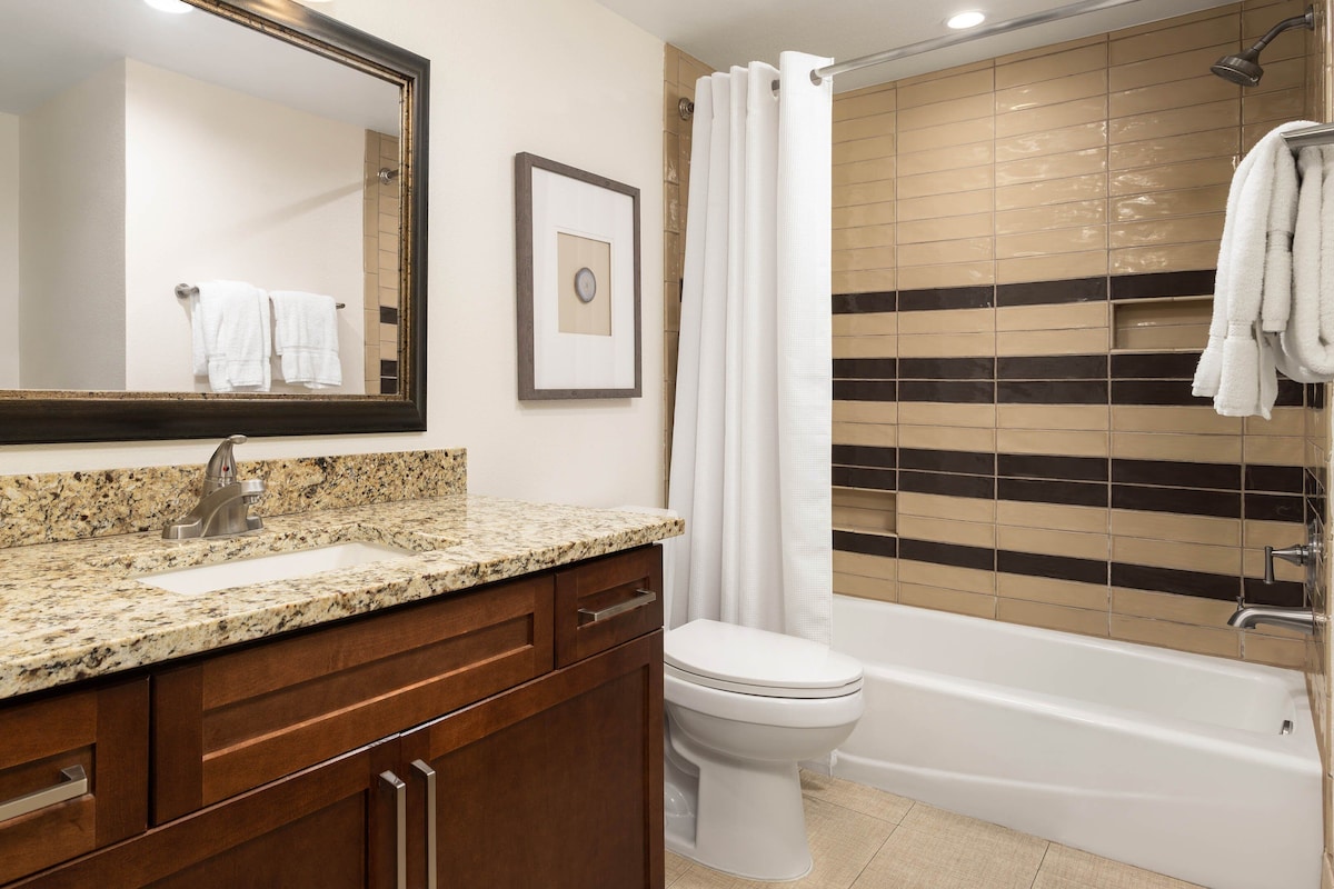 Une salle de bain avec un lavabo doté d’un plan de travail en granit, un miroir, des toilettes et une baignoire avec un rideau de douche.