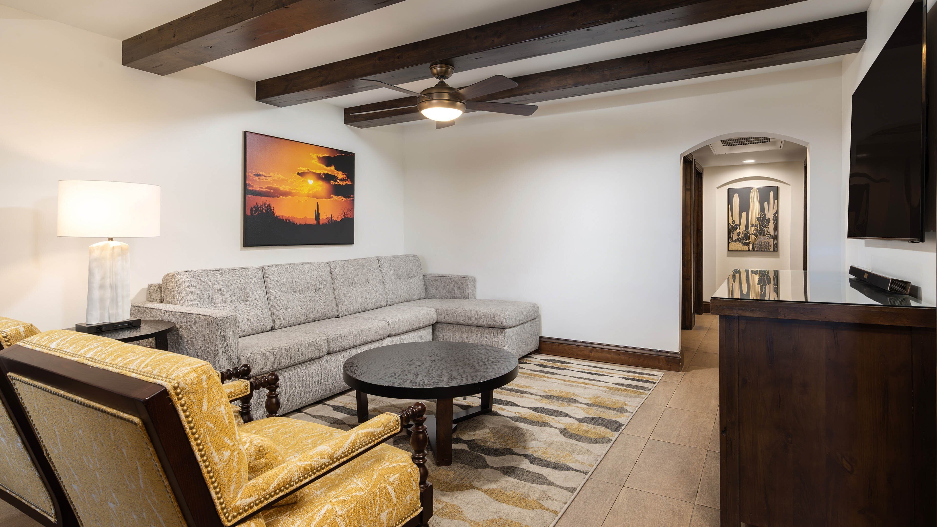 A living room with a sectional sofa, a round coffee table, a yellow armchair, a lamp, a ceiling fan, and artwork on the walls.
