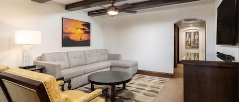 A living room with a sectional sofa, a round coffee table, a yellow armchair, a lamp, a ceiling fan, and artwork on the walls.