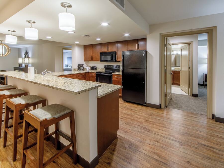 A modern kitchen with a central island, bar stools, wooden cabinets, a black refrigerator, and a microwave.