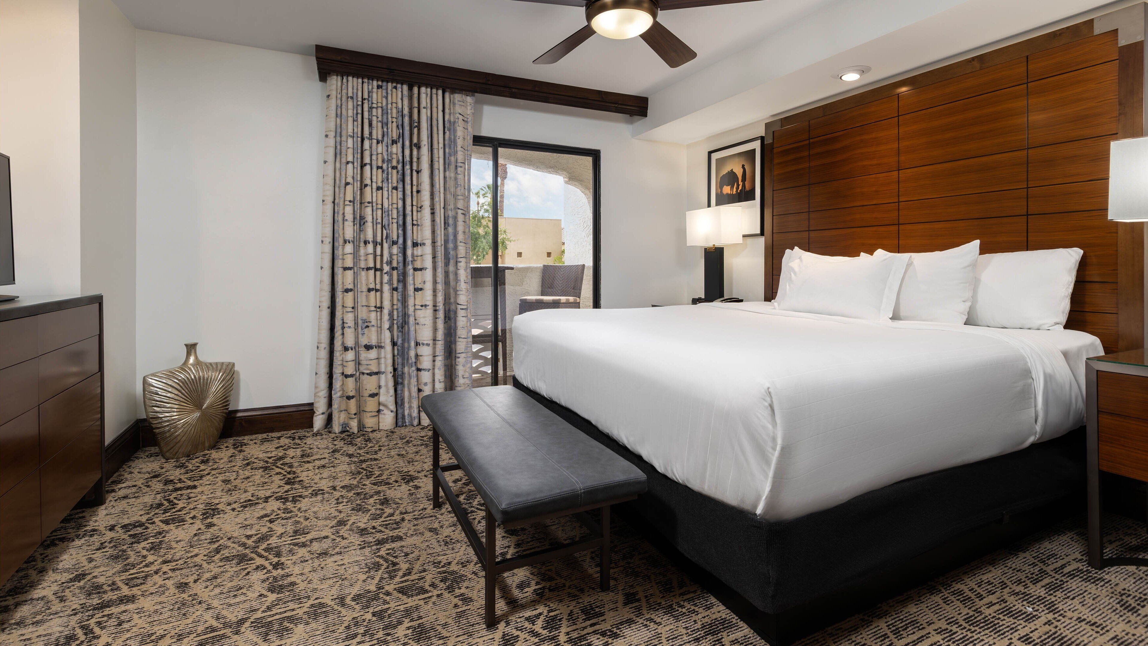 A hotel room with a large bed, a bench, a desk, a chair, and a ceiling fan.