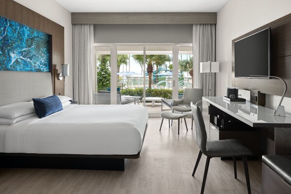 Ein modernes Hotelzimmer mit einem großen Bett, einem Schreibtisch, einem Stuhl, einem Fernseher und Blick auf einen Pool und Palmen.