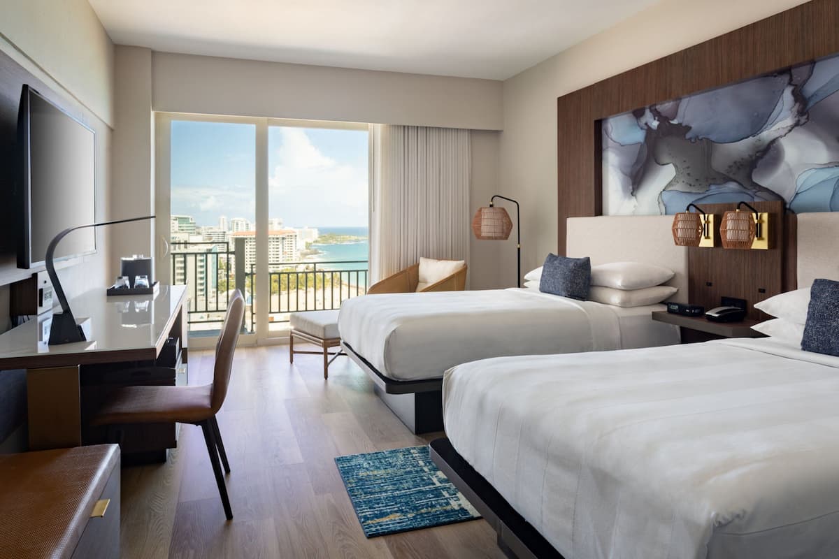 Ein Hotelzimmer mit zwei Betten, einem Schreibtisch, einem Sessel, einem Fernseher und einem großen Fenster mit Blick auf die Stadt.