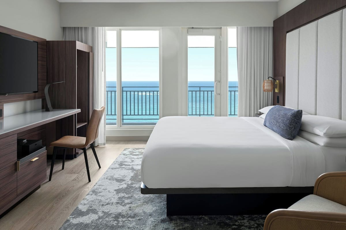 Ein modernes Hotelzimmer mit einem großen Bett, einem Schreibtisch mit Stuhl, einem Fernseher und einem Balkon mit Meerblick.