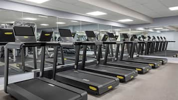 Una fila de cintas de correr en un gimnasio con paredes de espejo e iluminación en el techo.