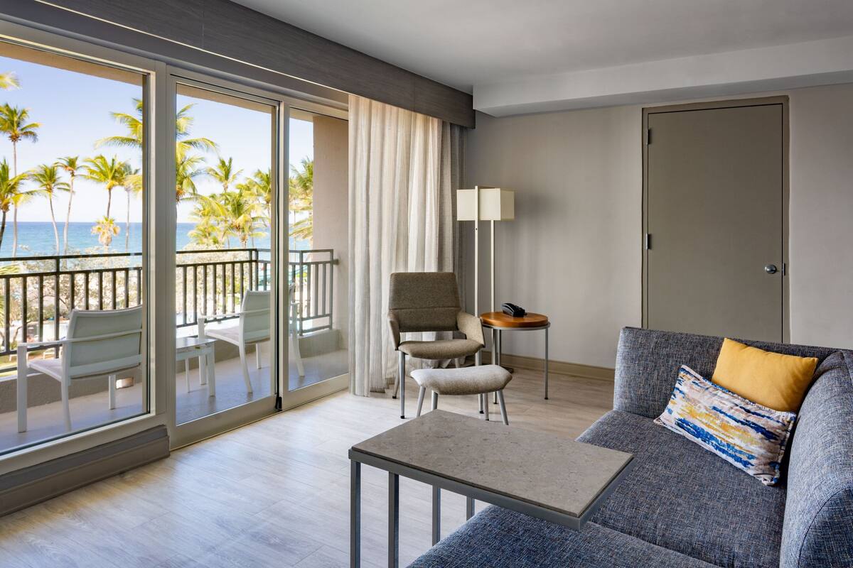 Ein modernes Hotelzimmer mit Balkon, einer Couch, einem Sessel, einem kleinen Tisch und einer Lampe.