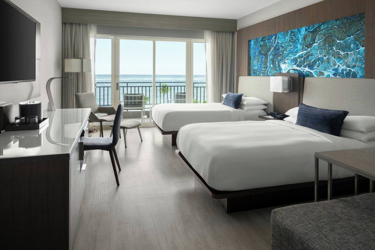 Ein Hotelzimmer mit zwei Betten, einem Schreibtisch, einem Stuhl, einem Fernseher und einem großen Fenster mit Meerblick.