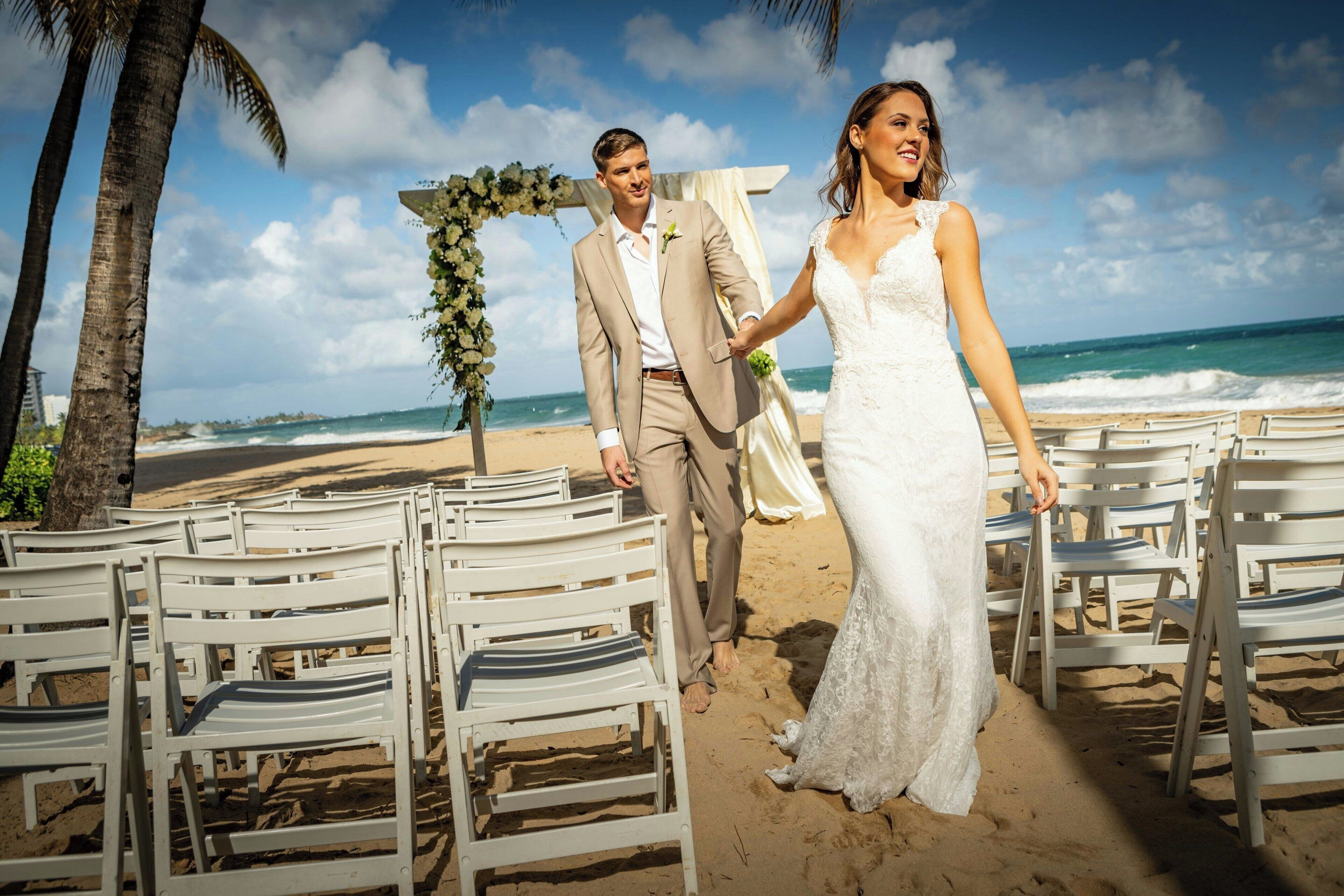 Une cérémonie de mariage sur la plage, avec des chaises blanches disposées en rangées, une arche fleurie et une vue dégagée sur l’océan.