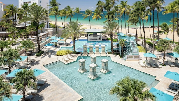 A resort with a central pool surrounded by palm trees and lounge chairs, a beach in the background, and a tall building to the left. - San Juan Marriott Resort and Stellaris Casino (San Juan)