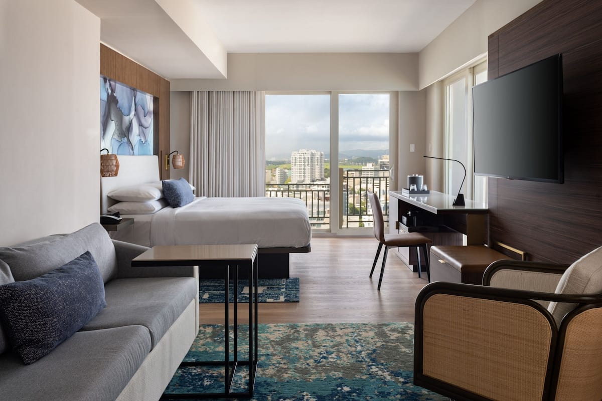 Ein modernes Hotelzimmer mit Bett, einer Sitzecke, einem Schreibtisch und einem Fernseher. Das Zimmer verfügt über ein großes Fenster mit Blick auf die Stadt.