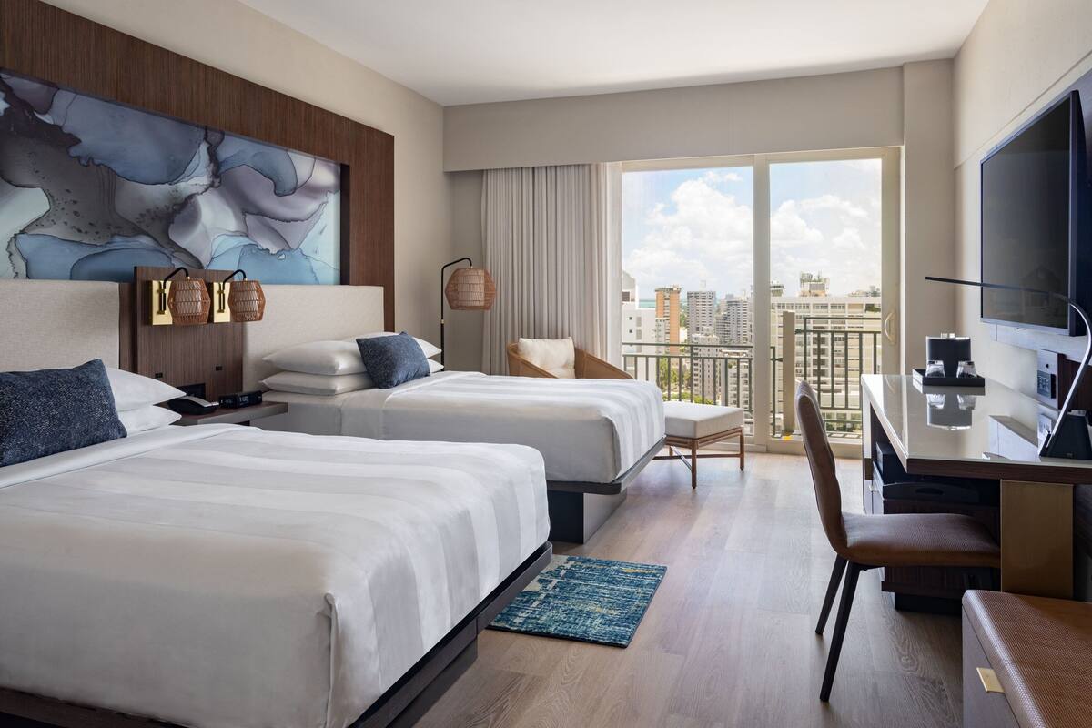 Ein Hotelzimmer mit zwei Betten, einem Schreibtisch, einem Stuhl, einem Fernseher und einem großen Fenster mit Blick auf die Stadt.