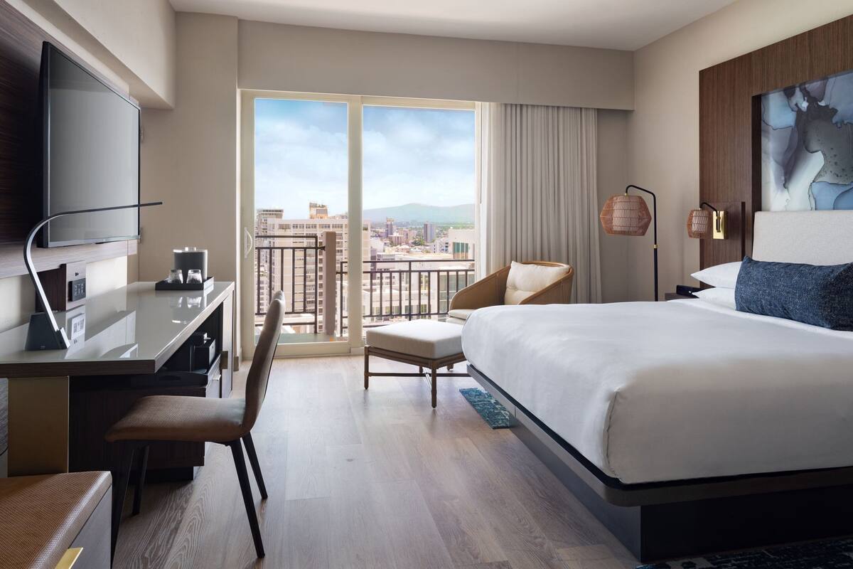 Ein Hotelzimmer mit Bett, Schreibtisch, Stuhl, Fernseher und einem großen Fenster mit Blick auf die Stadt.