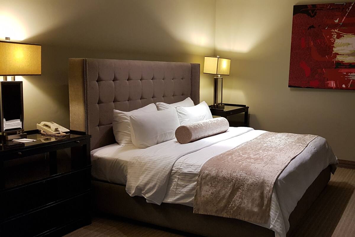 A neatly made bed with a tufted headboard, two bedside lamps, a telephone, and a large abstract painting on the wall.
