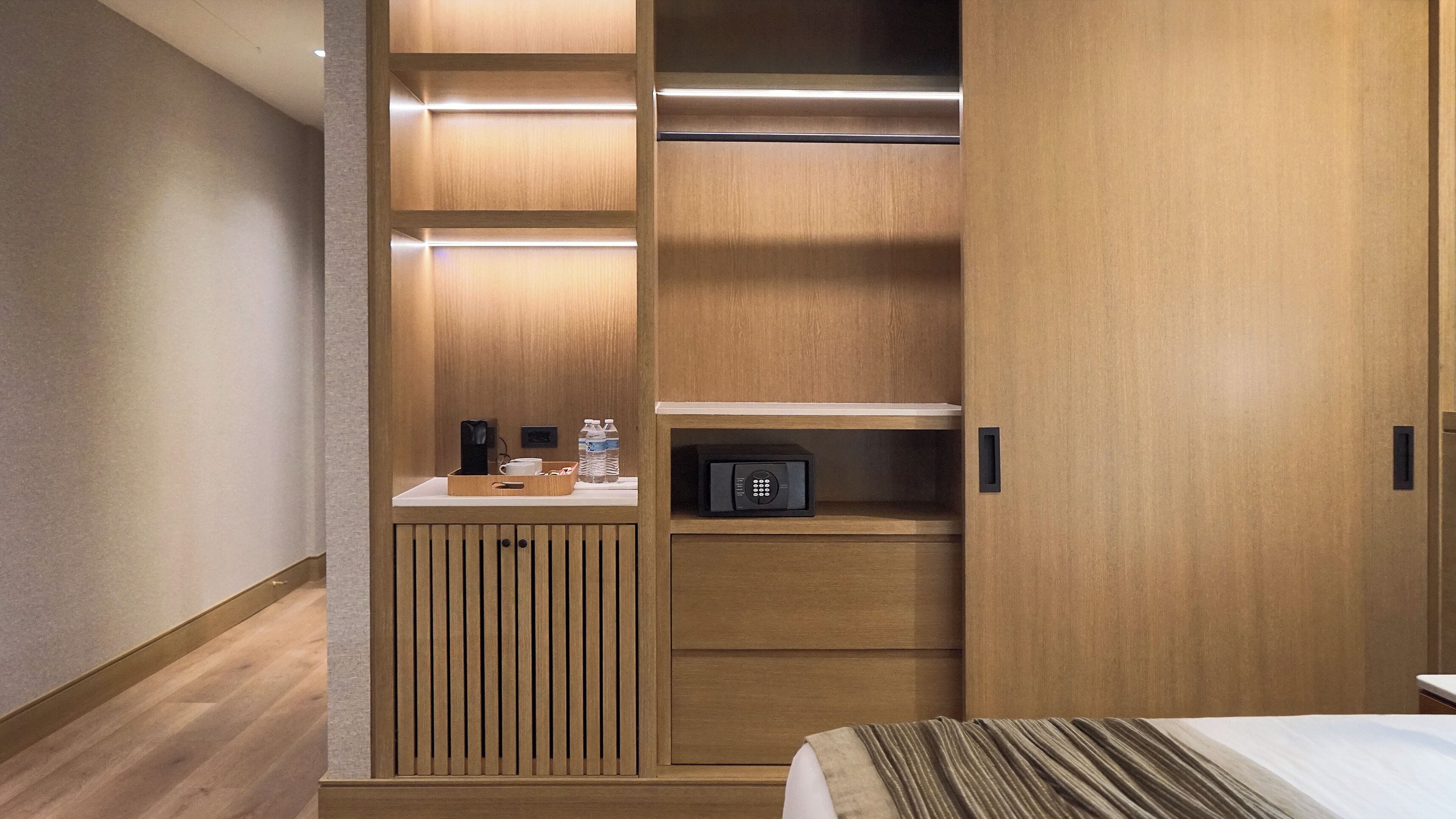 Una habitación de hotel con un armario de madera, una mesita de noche con lámpara y una cama con manta a rayas.