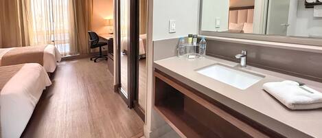 A hotel room with two beds, a desk, a chair, a large mirror, a bathroom with a sink, and toiletries.