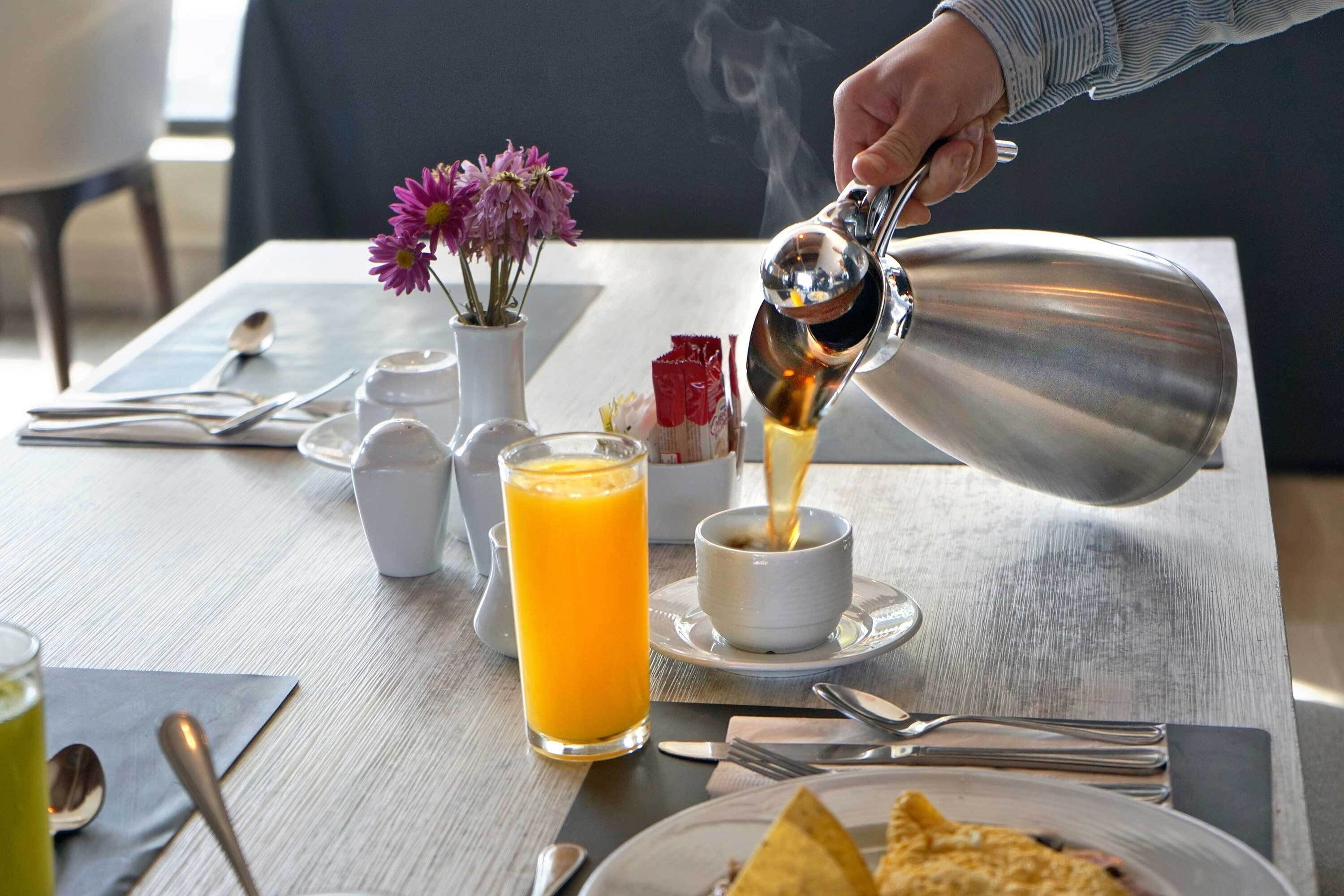 A breakfast setting with a person pouring a hot beverage into a cup, a glass of orange juice, and a plate with food.