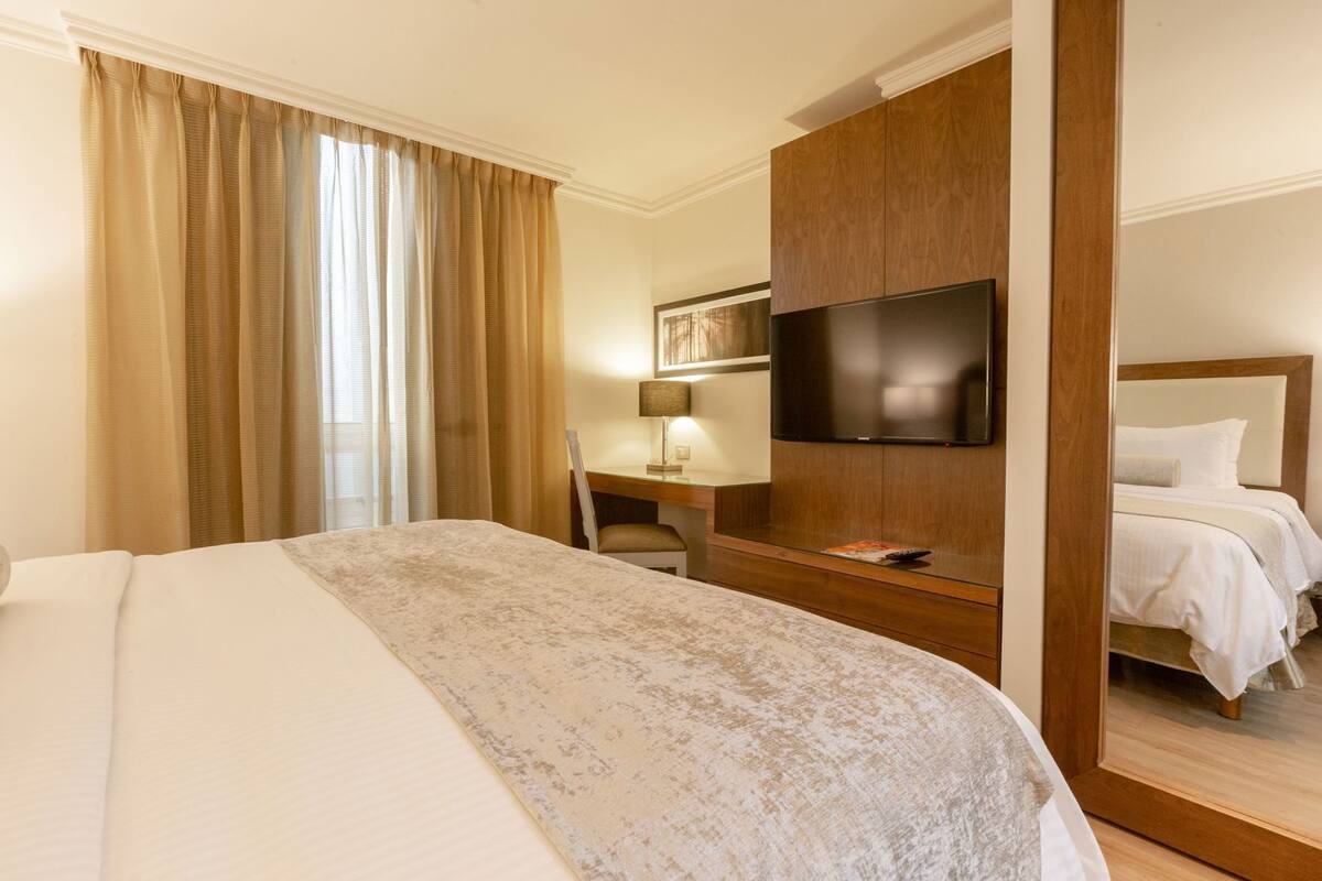 A hotel room with a bed, a desk, a TV, and a second bed.