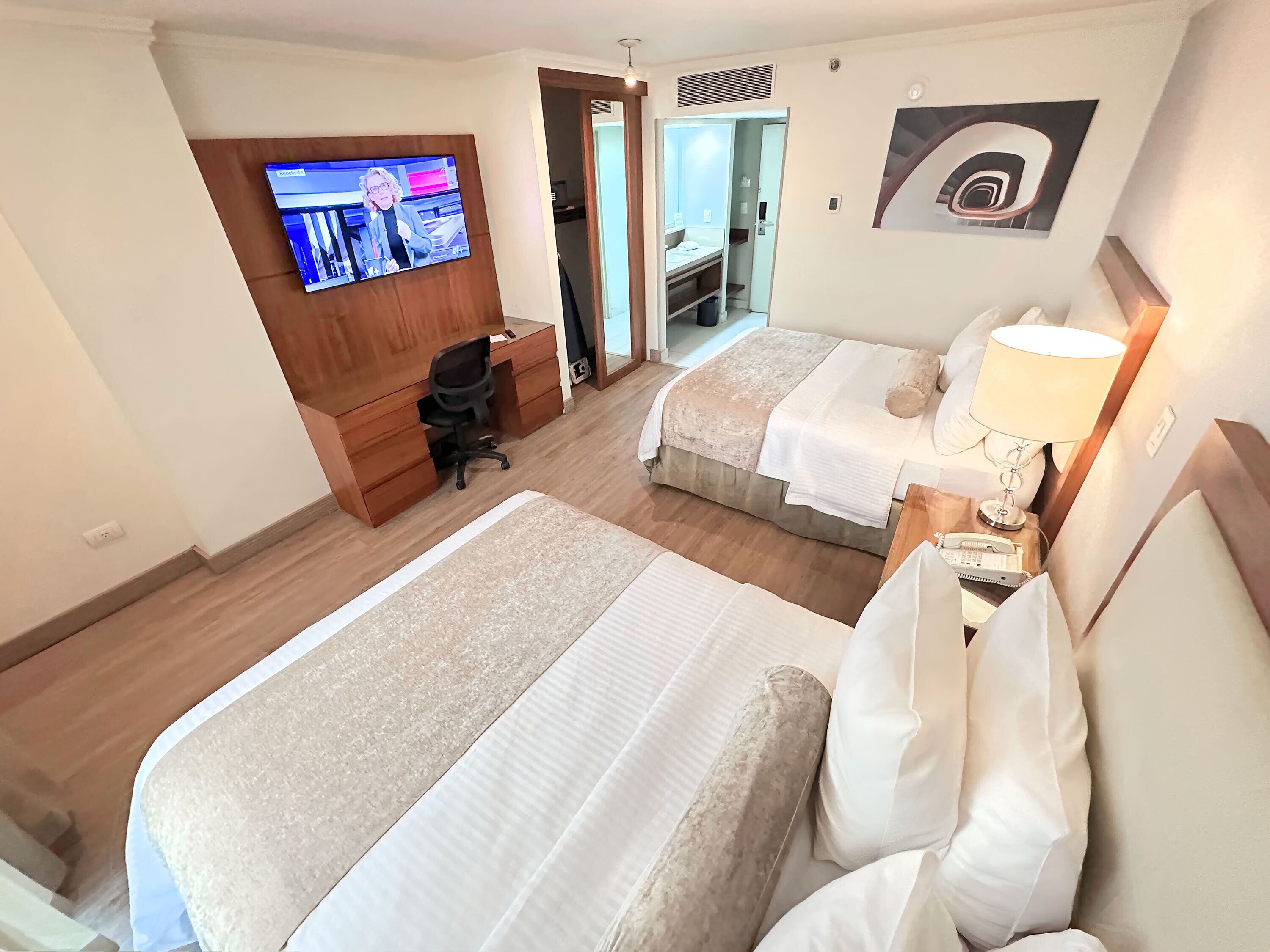 Habitación de hotel con dos camas, un escritorio, televisión y baño visible a través de una puerta abierta.