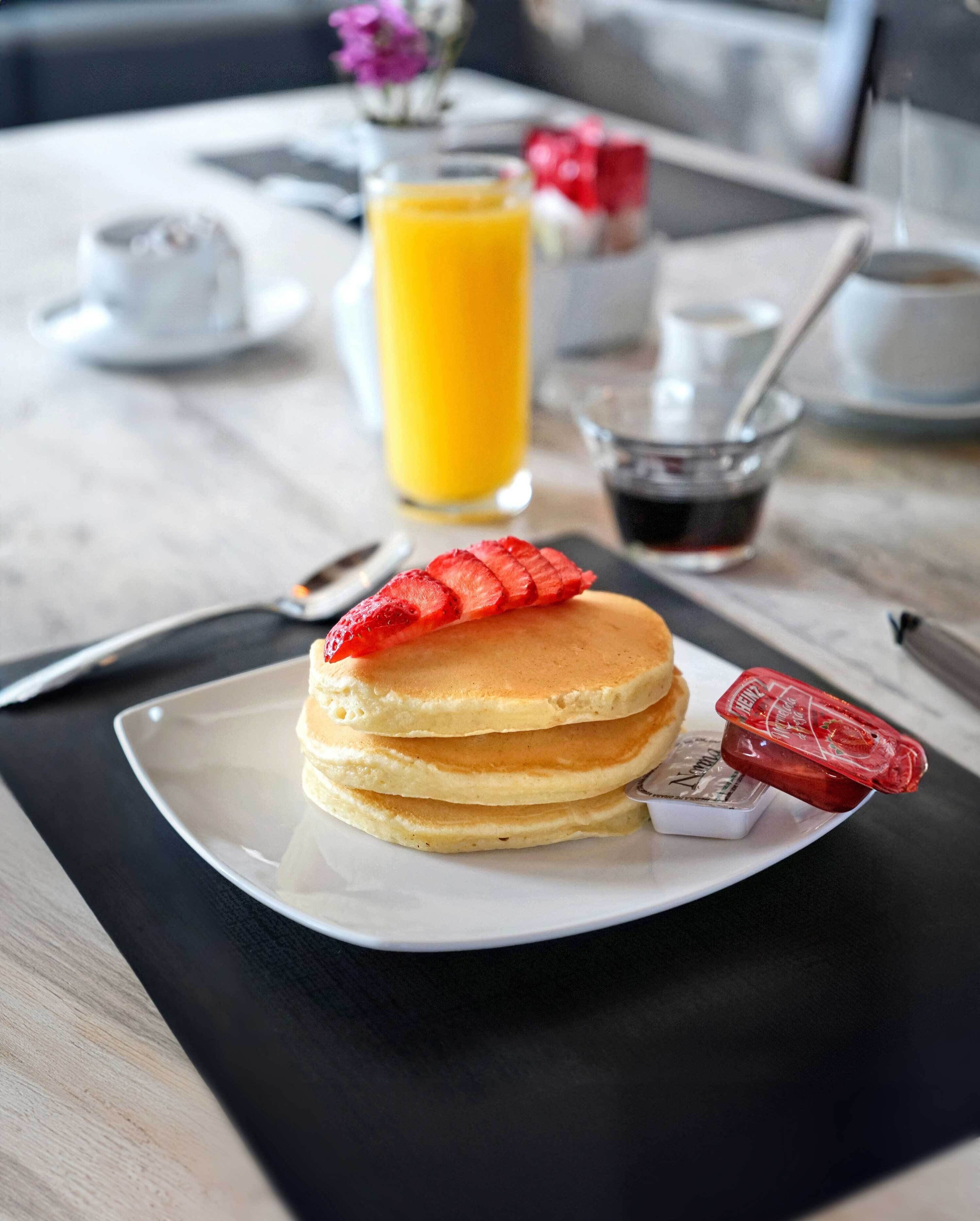 Une table dressée pour le petit-déjeuner avec des crêpes, un verre de jus d’orange, une tasse de café et divers condiments.