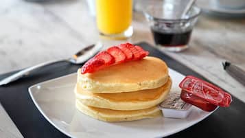 Une table dressée pour le petit-déjeuner avec des crêpes, un verre de jus d’orange, une tasse de café et divers condiments.