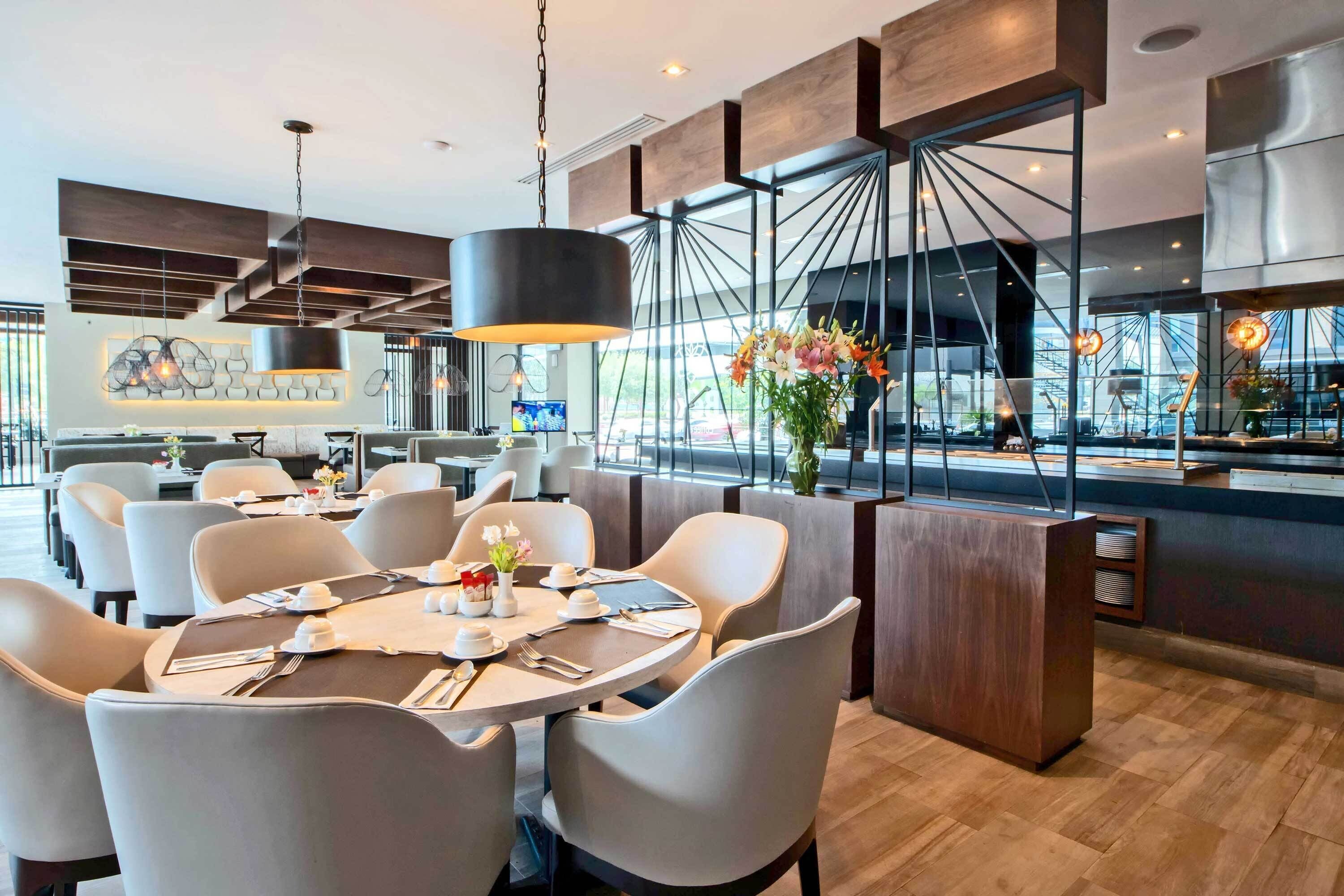 Interior moderno de restaurante com área de refeições central, tetos altos e vista da cidade através de grandes janelas.