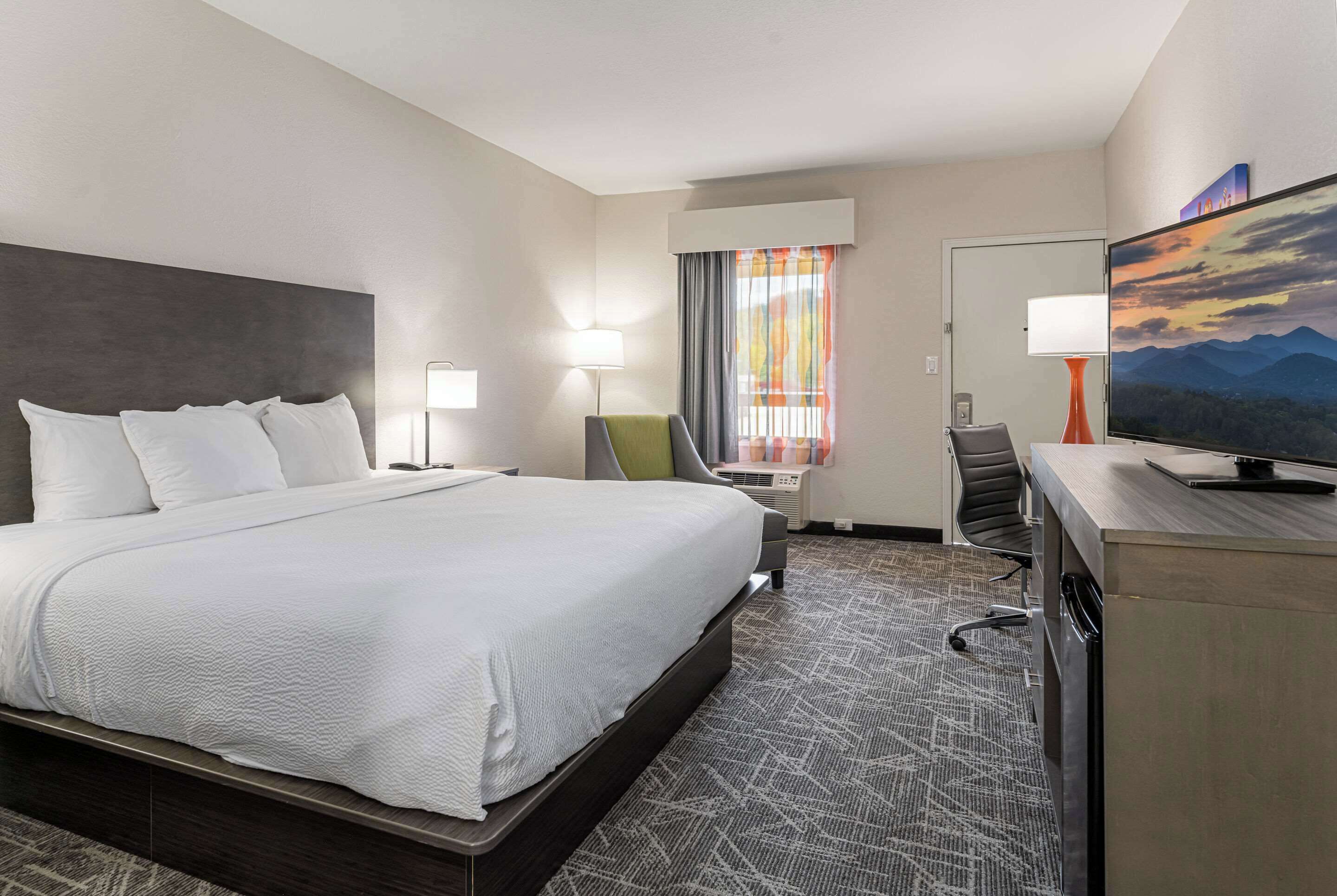 A hotel room with a large bed, a desk with a chair, a television, and a window with curtains.