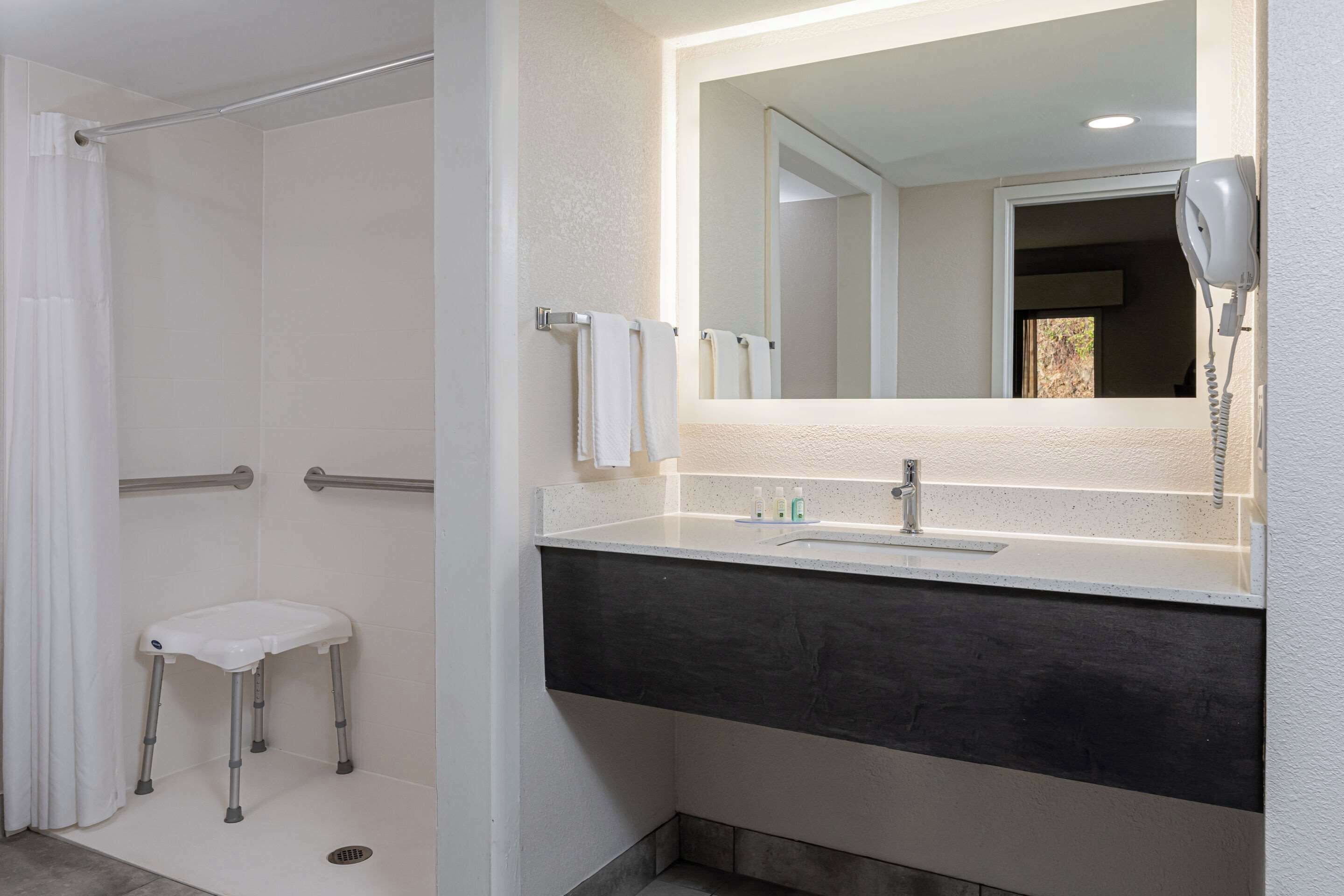 A bathroom with a shower, a white toilet, a vanity with a mirror, and a hairdryer mounted on the wall.
