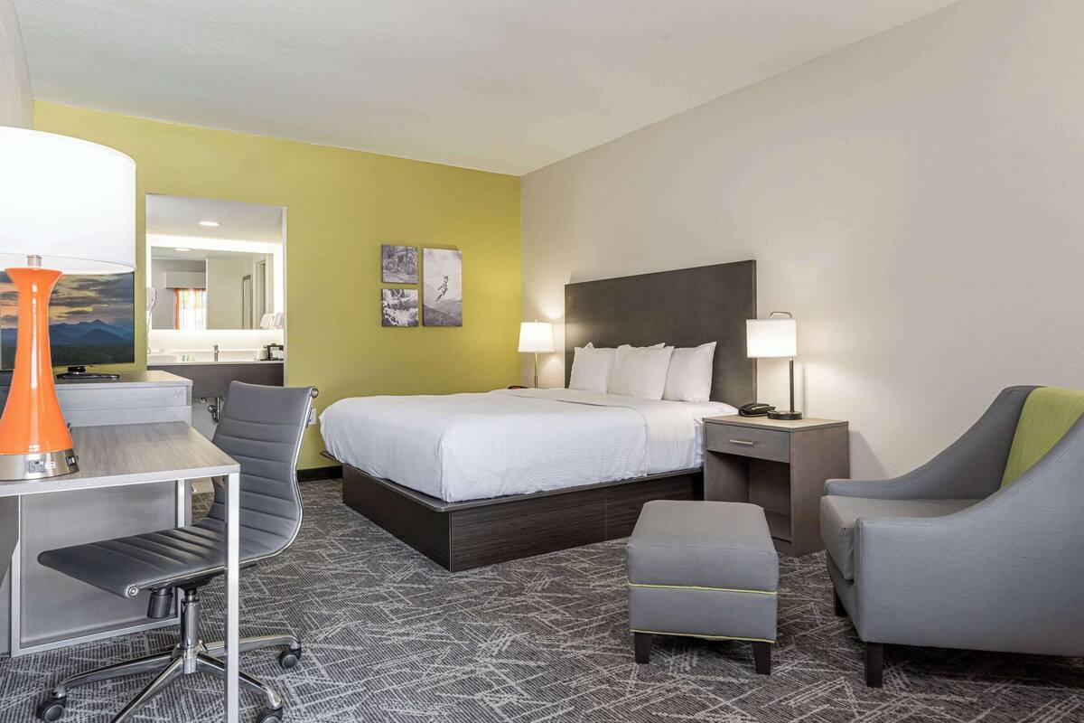 A hotel room with a bed, two bedside tables, a desk with a chair, a small ottoman, and a desk lamp.