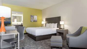 A hotel room with a bed, two bedside tables, a desk with a chair, a small ottoman, and a desk lamp.