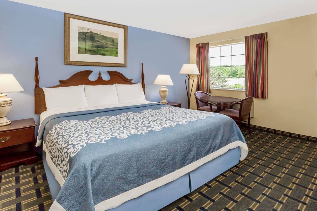 A hotel room with a large bed, two bedside tables with lamps, a chair, a window with curtains, and a framed picture on the wall.