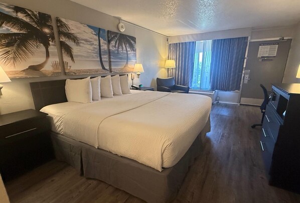 Habitación de hotel con una cama grande, dos mesitas de noche, un escritorio y una silla. Se ve un ventanal con cortinas, un cuadro de palmeras y un letrero en la pared.