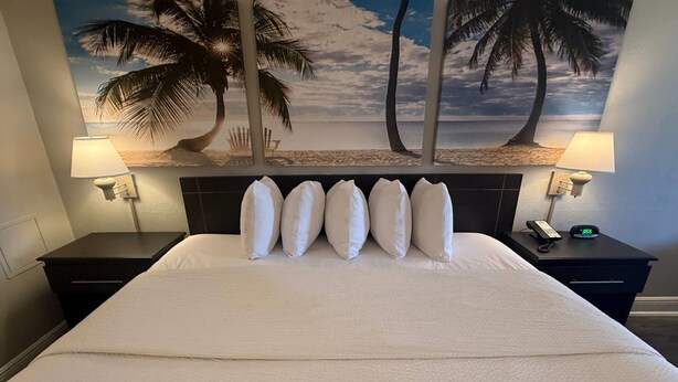 Une chambre d’hôtel avec un grand lit, des oreillers blancs, une tête de lit, deux tables de chevet avec des lampes, un téléphone et une fresque murale représentant des palmiers et une plage.