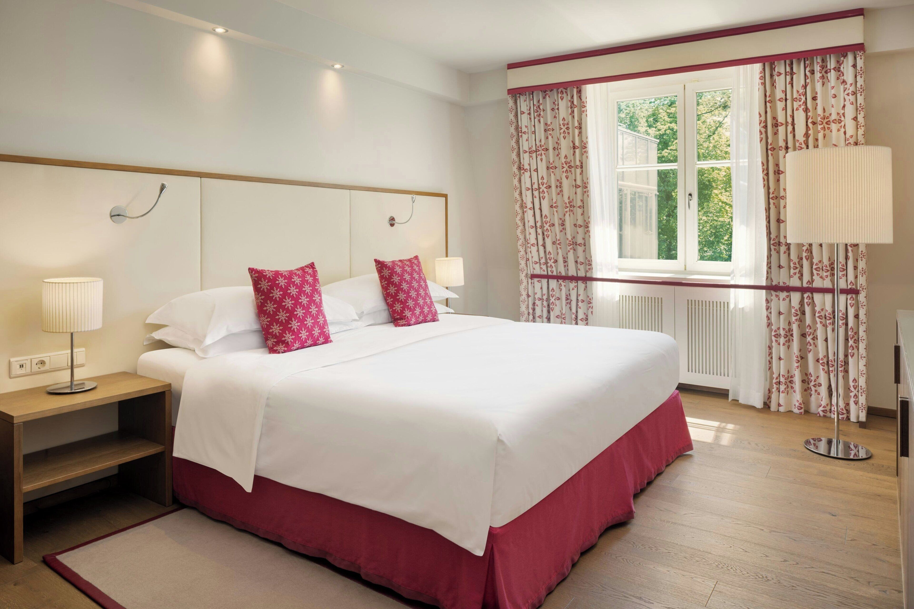 A hotel room with a large bed, two bedside tables with lamps, a window with floral curtains, and a radiator under the window.