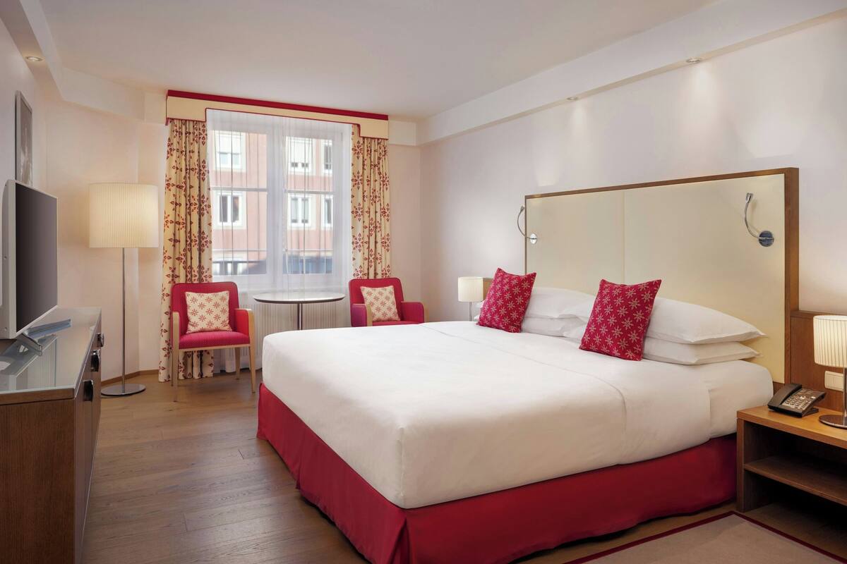 Habitación de hotel con una cama grande, un escritorio, una silla, una mesita, un espejo y una ventana con cortinas.